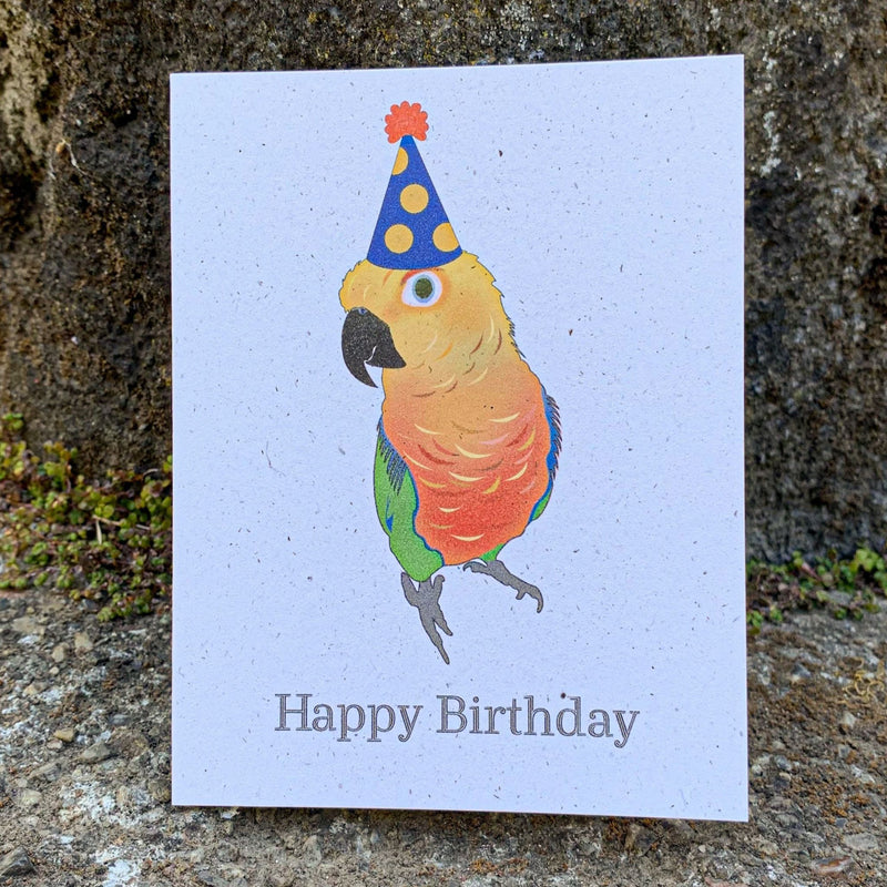 A cream colored speckled greeting card, with Happy Birthday written at the bottom in black ink. Centered above the text is a curious jenday conure with shades of yellow, orange, red, green, and blue, wearing a dotted gold and blue hat with an orange poof.