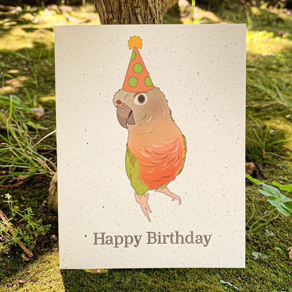 A cream colored speckled greeting card, with Happy Birthday written at the bottom in black ink. Centered above the text is an inquisitive pineapple green cheek conure with shades of orange, grey, tan, and green, wearing a dotted light green and orange hat with a golden poof.