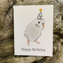 A cream colored speckled greeting card, with Happy Birthday written at the bottom in black ink. Centered above the text is a fluffy white and grey parakeet, wearing a dotted white and dark grey hat with a golden poof.