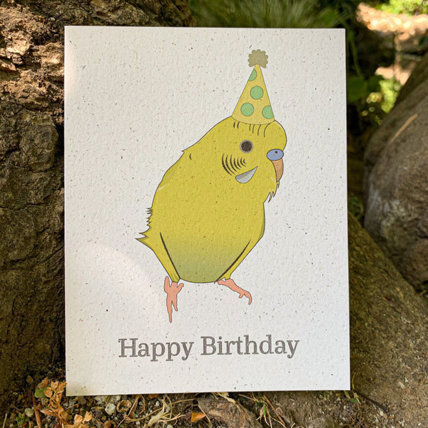 A cream colored speckled greeting card, with Happy Birthday written at the bottom in black ink. Centered above the text is a fluffy yellow and green colored parakeet, wearing a dotted gold and light green colored hat with a tan colored poof.