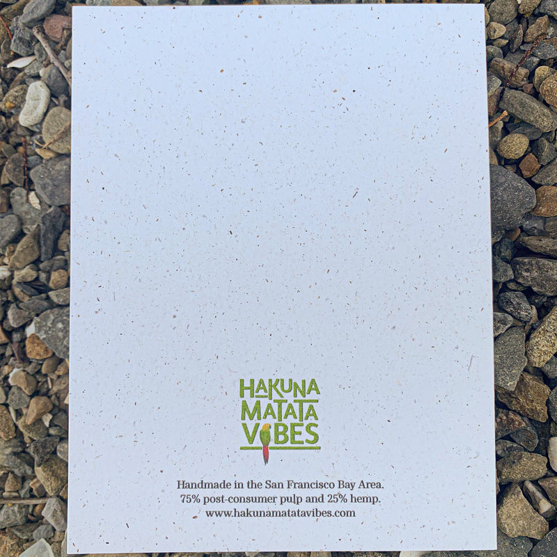 Centered about a third of the way down on the back of the cream colored speckled greeting card, is the Hakuna Matata Vibes logo with each word written out vertically in light green. The black text centered right below says, "Handmade in the San Francisco Bay Area. 75% post-consumer pulp and 25% hemp, www. hakunamatatavibes.com